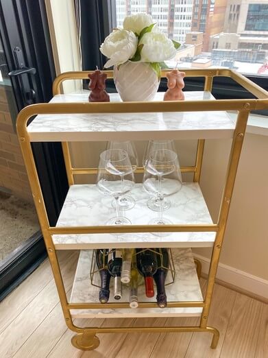 Bar wagon with 3 shelves. On the top shelf is a white vase with white fake flower and body candless.4 glasses sit on the middle shelf. and on the bottom shelf there is a unique shaped wine glass.
